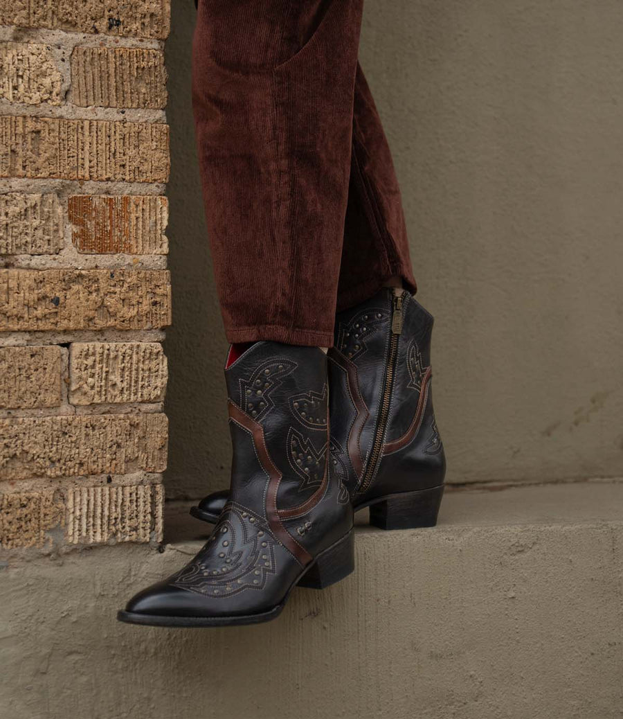 Close-up of Chamise Rustic Western Cowboy Boots in Black Teak Rustic styled outdoors, highlighting handcrafted details and rustic western charm for bohemian fashion outfits